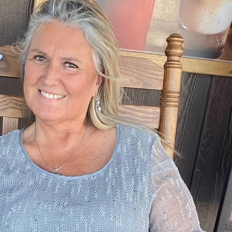Smiling woman with silver hair in a sparkly top sitting on a wooden chair.
