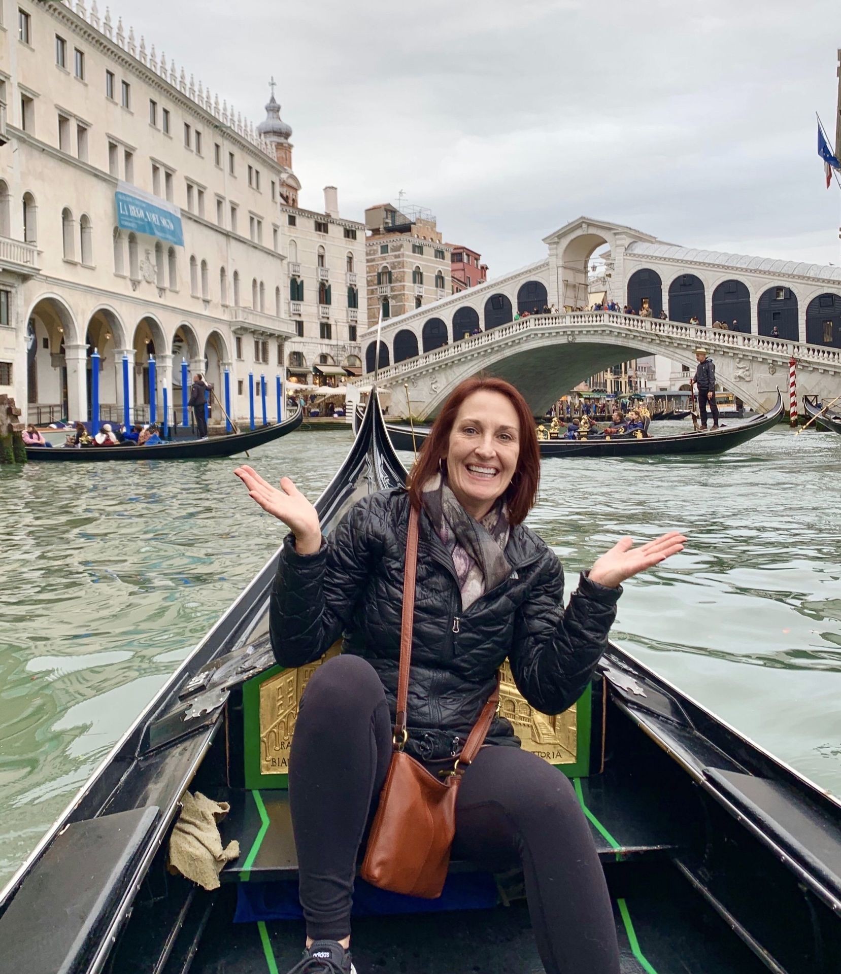 Person sitting on a gondola in Venice, smiling with the Rialto Bridge in the background.
