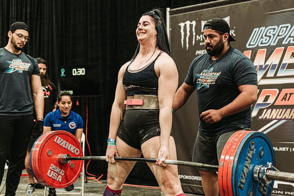 Woman performing a deadlift at a weightlifting competition with coaches and spectators nearby.