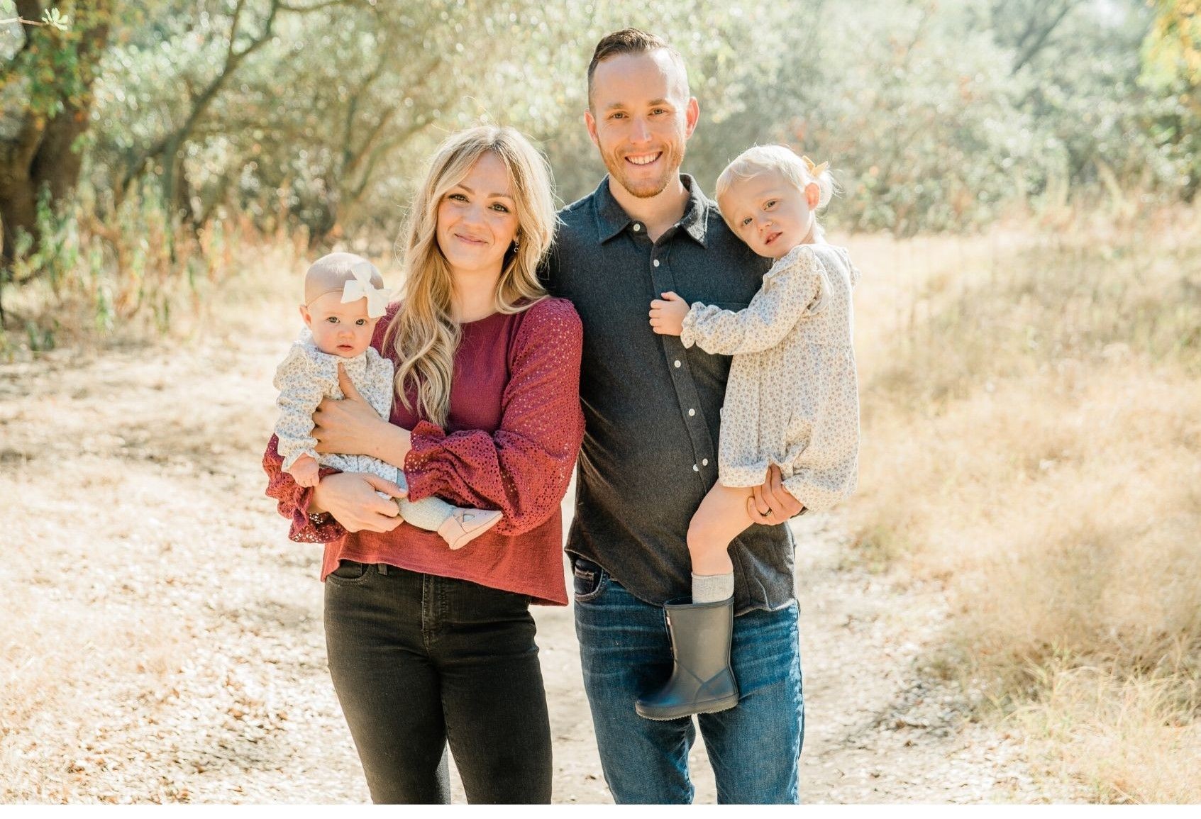 Smiling couple with two young children standing on a wooded path during a sunny day.