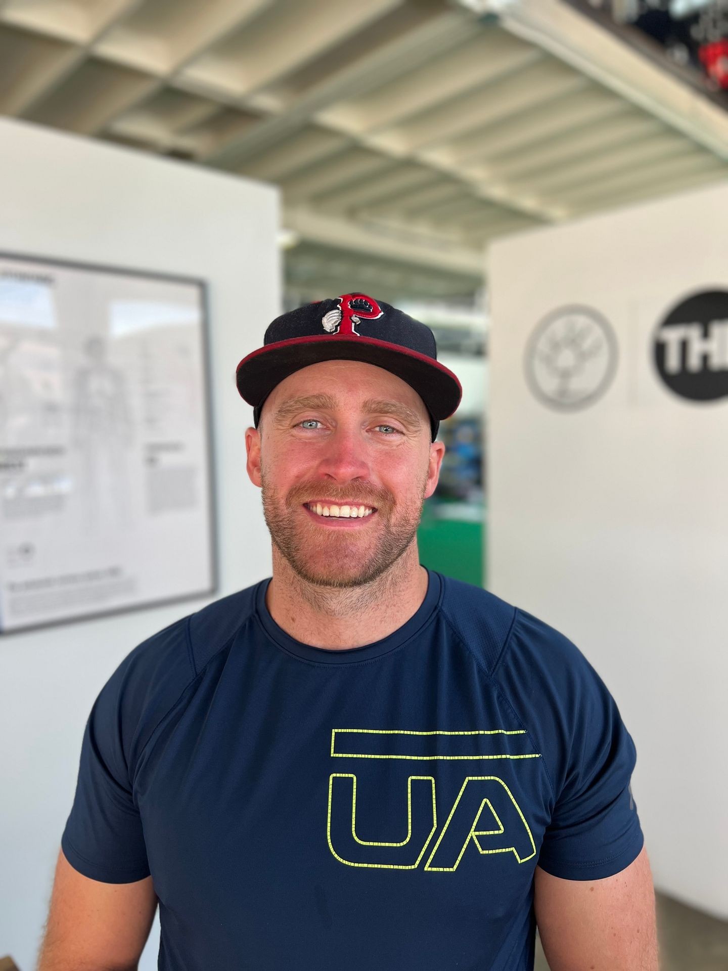 Smiling man wearing a black cap and blue UA t-shirt in a bright indoor setting.